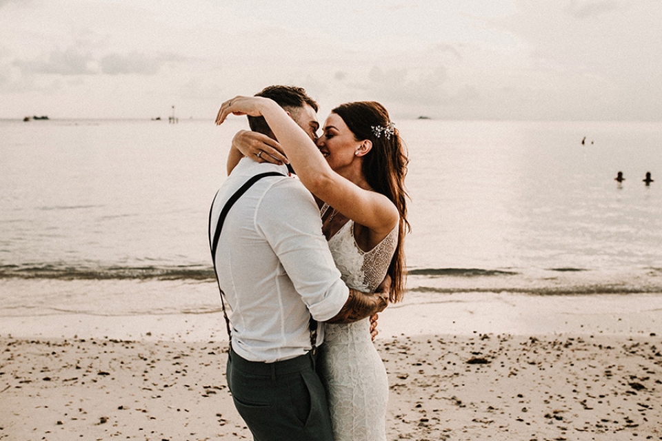bride flings arms around her husband neck as they kiss on the beach